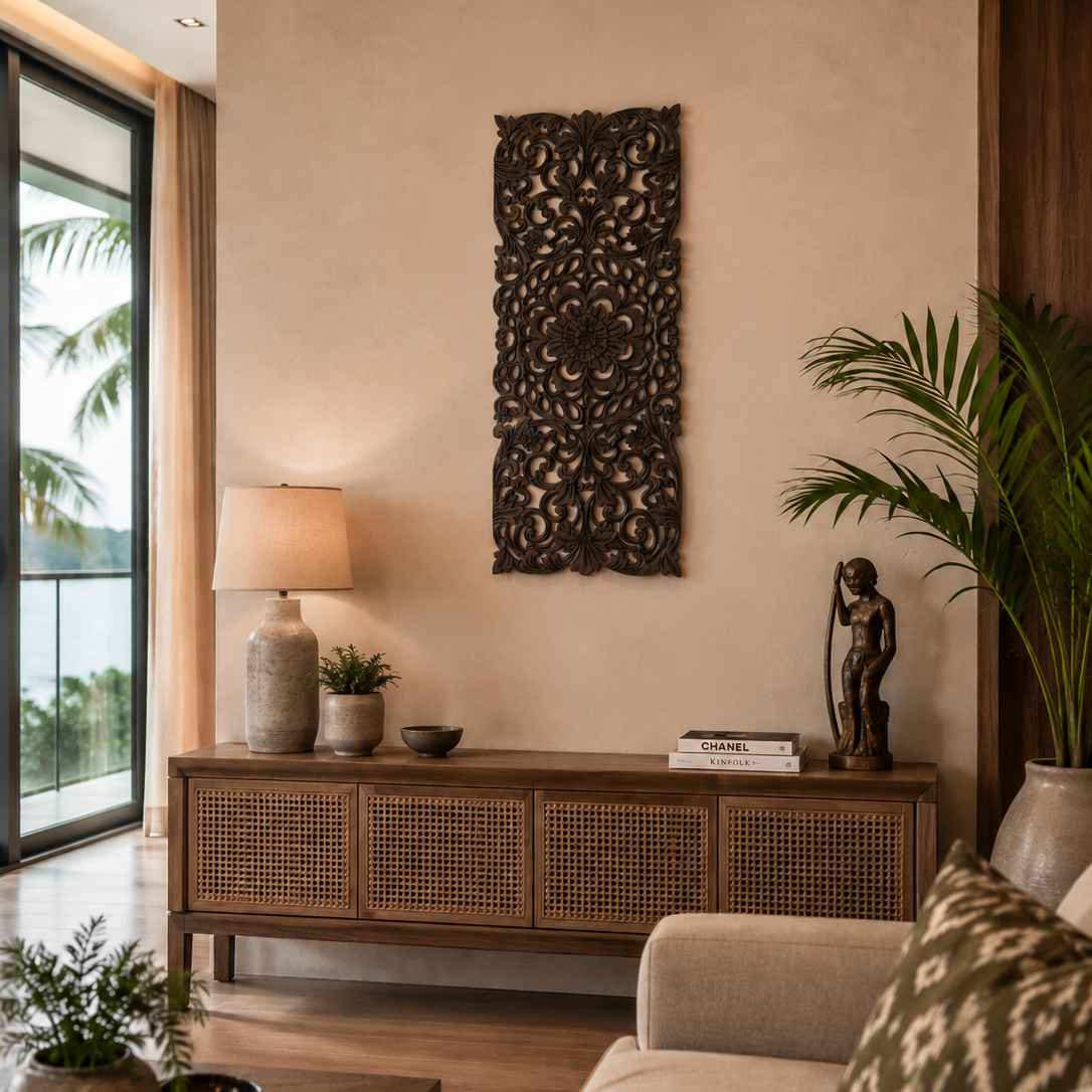A styled interior scene showing a beige wall with a tall dark brown carved wall hanging, a wooden console cabinet with woven cane doors, a table lamp, books, a small statue, and a large potted palm. Part of a sofa and a patterned pillow are visible in the foreground.